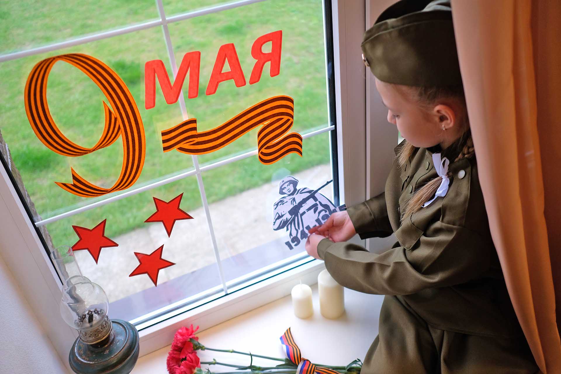 Фотография девочка в военной форме украшает окно к 9 Мая.